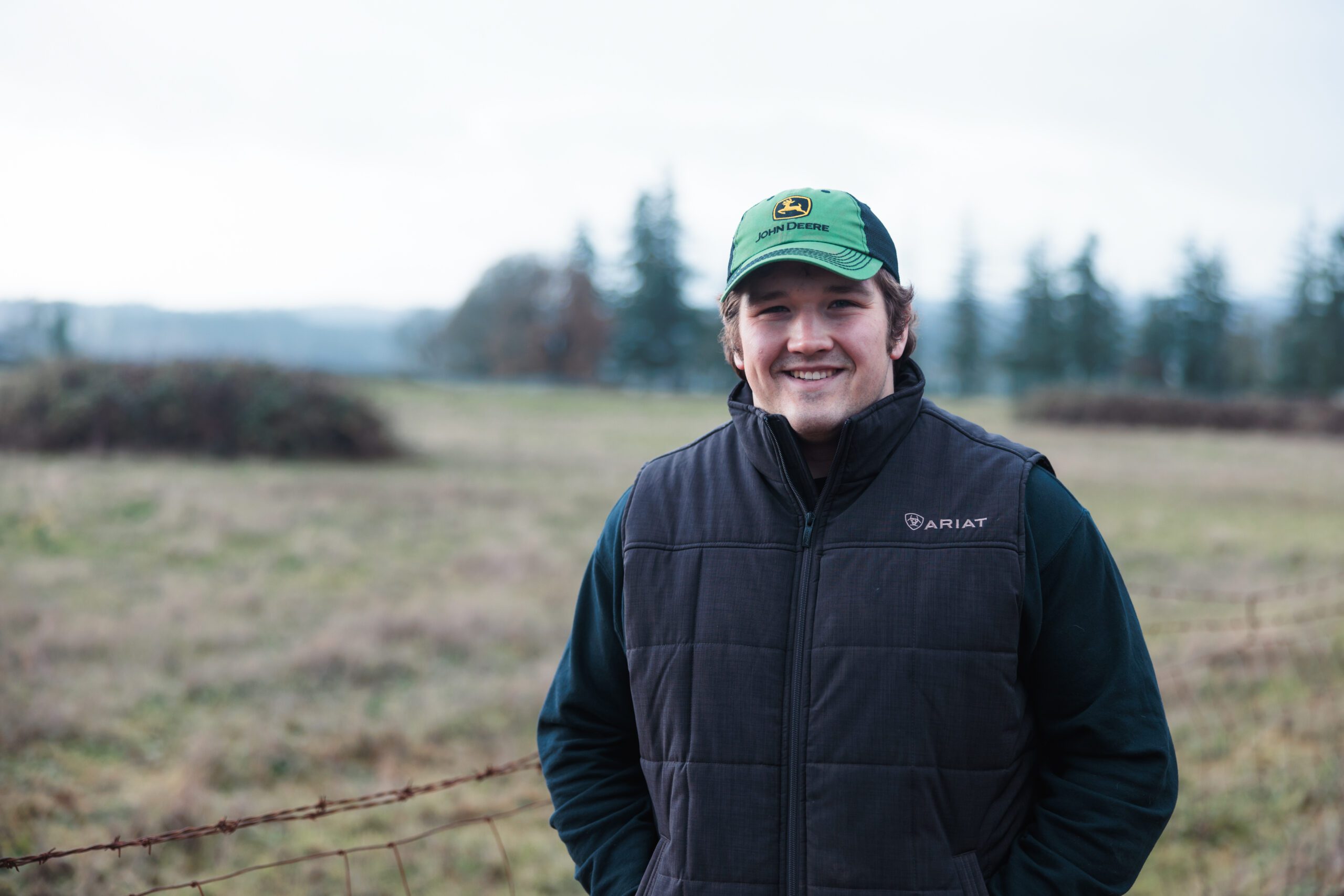 A person wearing a green cap and dark vest smiles while standing in a grassy field with trees in the background. The scene appears outdoors and cloudy.