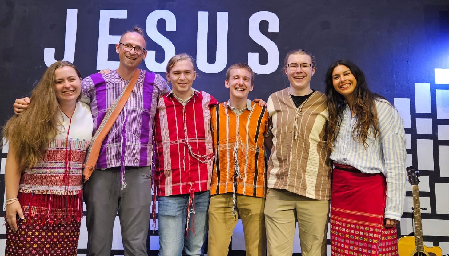 A group of six smiling people stand side by side, wearing colorful traditional outfits. They are positioned in front of a wall with the word JESUS displayed prominently at the top. A guitar is partially visible on the right side.