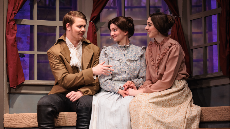 Three actors in period costumes sit on a window seat with red curtains. The man on the left gestures while the two women on the right listen intently, creating a scene suggestive of a theatrical performance or historical drama.